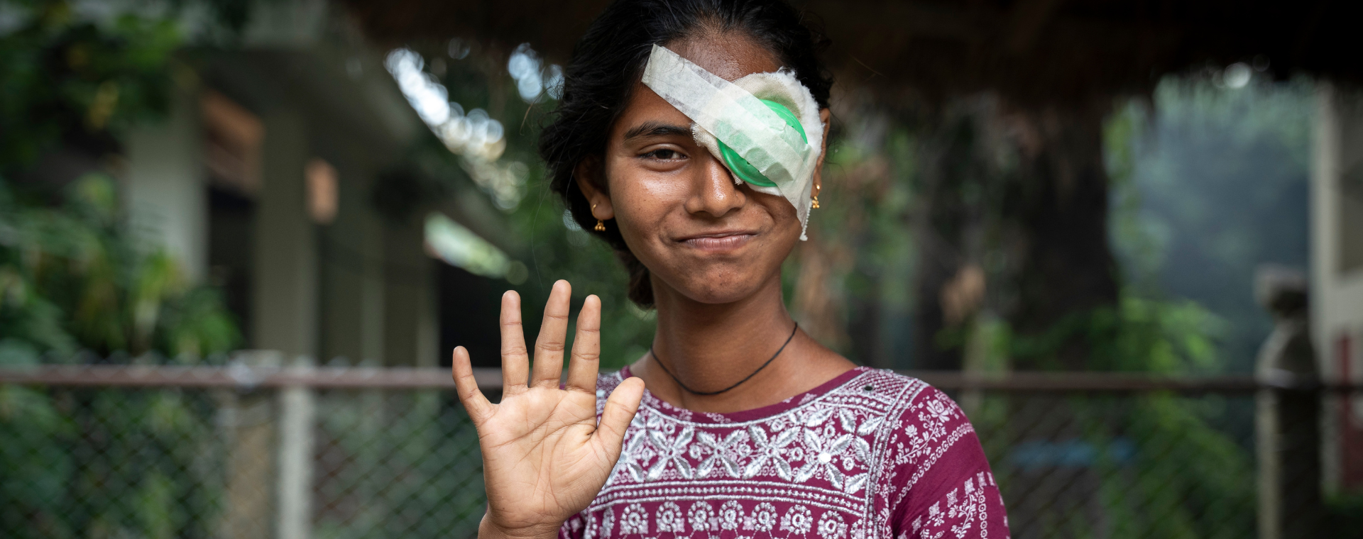 A girl with a green eye patch on her left eye waves and smiles at the camera following eye surgery. A donation this Christmas to CBM UK will ensure more children receive the eye care and treatment they need to prevent blindness.