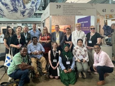 A group of people at COP 30 smile at the camera. Behind them is a booth in a conference hall with various disability organisations listed.