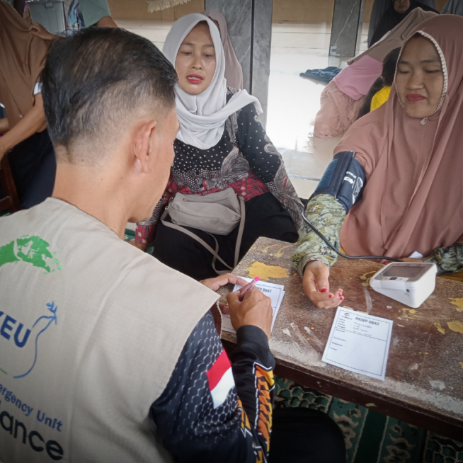 A man, wearing a Yakkum Emergency Unit tabbard takes a blood pressure reading of a woman sat across the table from him.
