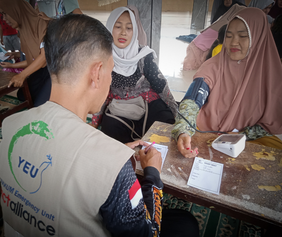 A man, wearing a Yakkum Emergency Unit tabbard takes a blood pressure reading of a woman sat across the table from him.