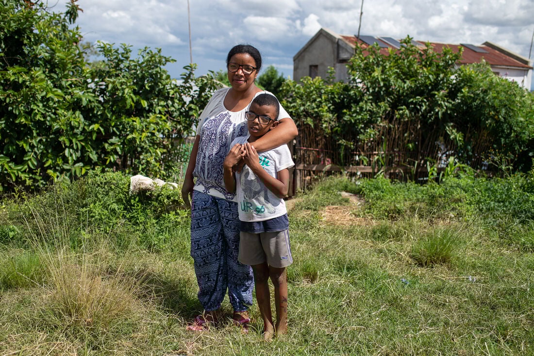 A middle aged Madagascan women wearing blue and white patterned cotton trousers and tshirt standing with her young son aged 10 wearing faded shorts and white tshirt and corrective black glasses. The woman has her arm around her son and they are standing in a garden with hedges in the background and on grass in their bare feet