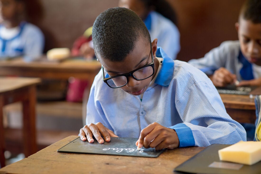Young Madagascan boy wearing a pale blue long sleeved shirt and black glasses writing with a piece of chalk on a chalkboard at a wooden desk in a classroom