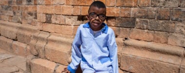 Boy aged 10 from Madagascar wearing a pale blue long sleeved shirt and black corrective sight glasses sitting beside a school building
