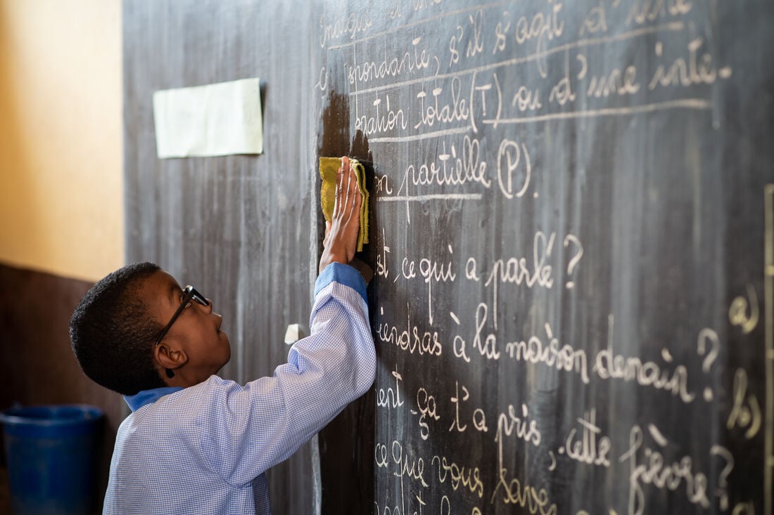 Young Madagascan boy aged 10 in a pale blue long sleeved shirt and wearing black corrective sight glasses writing on a blackboard with a piece of chalk