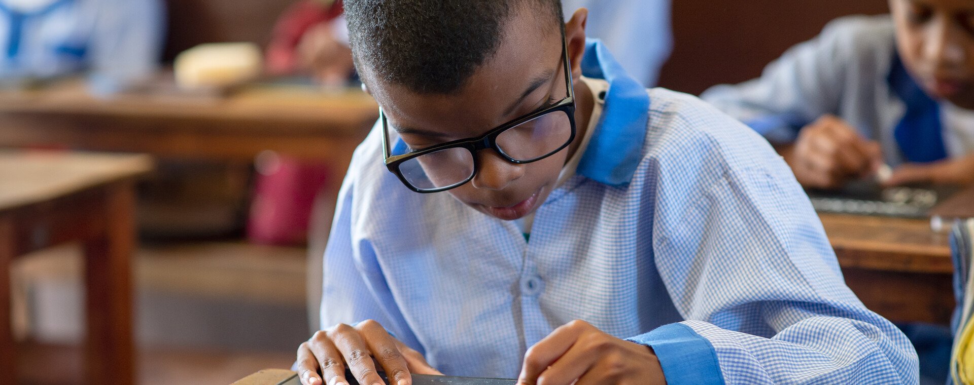 A boy wearing glasses focuses on writing in a school classroom.