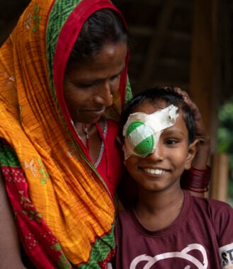 A woman wearing an orange, red and green sari, with her hand resting on her young son's head. He has a green bandage on his right eye, and they are both smiling.