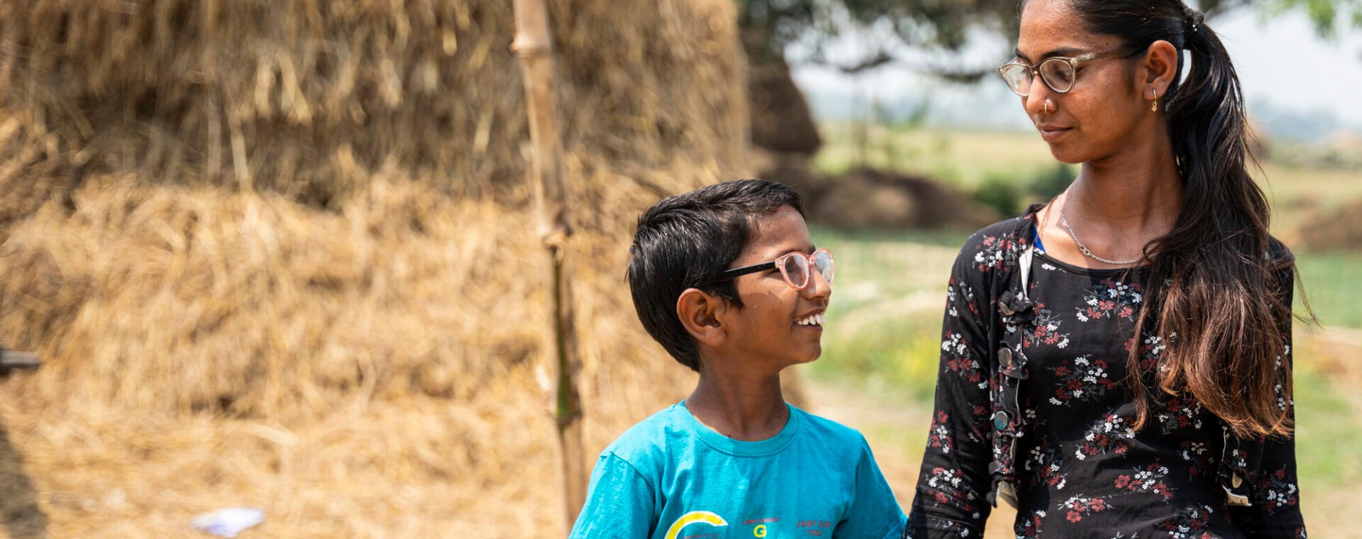 A young boy and girl holding hands and smiling at each other. She is wearing a floral black dress and he is wearing a blue tshirt and looking up t her.
