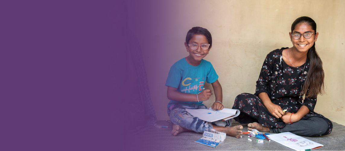 A Nepalese boy and girl sat cross-legged with exercise books and pens, smiling.