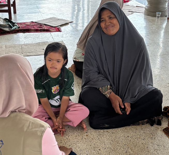 A little girl is pictured sitting on the floor next to her mother while they talk to a woman pictured in the corner of the image. They are sitting in an evacuation shelter after Indonesia Tropical Cyclone Senyar.
