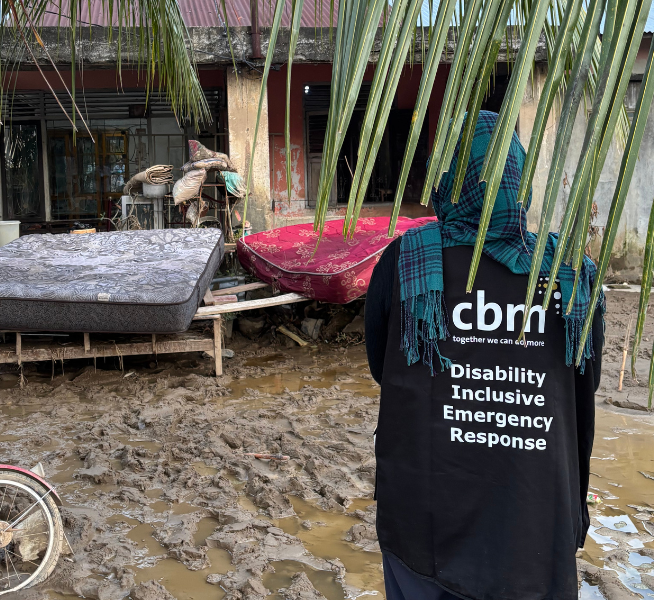 Indonesia Tropical Cyclone Senyar left homes destroyed. A home is pictured surrounded by mud and debris. A woman is standing facing the home wearing a black jacket with the writing 'CBM Disability Inclusive Emergency Response'.
