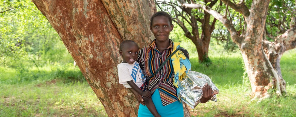 A woman holding a child on one hip and a bag of food supplement in the other hand stands smiling at the camera.