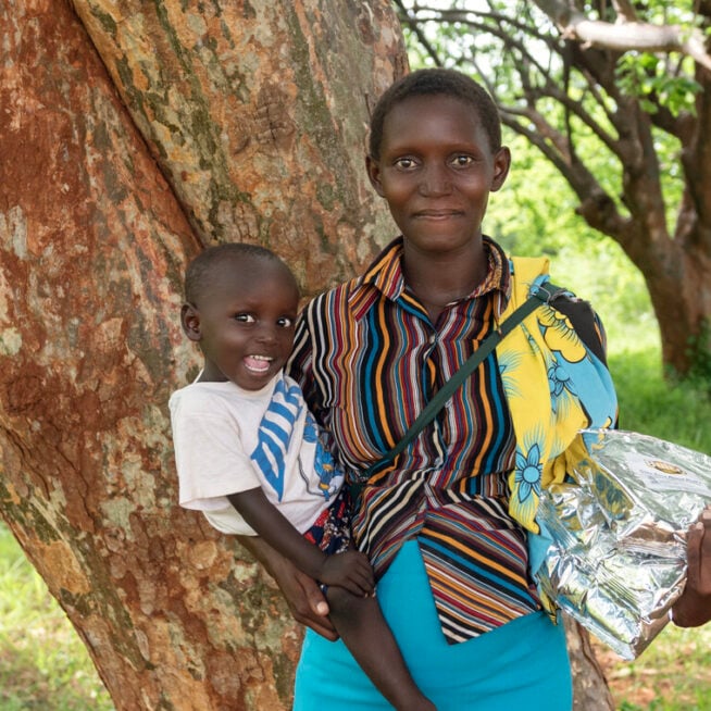 A woman holding a child on one hip and a bag of food supplement in the other hand stands smiling at the camera.