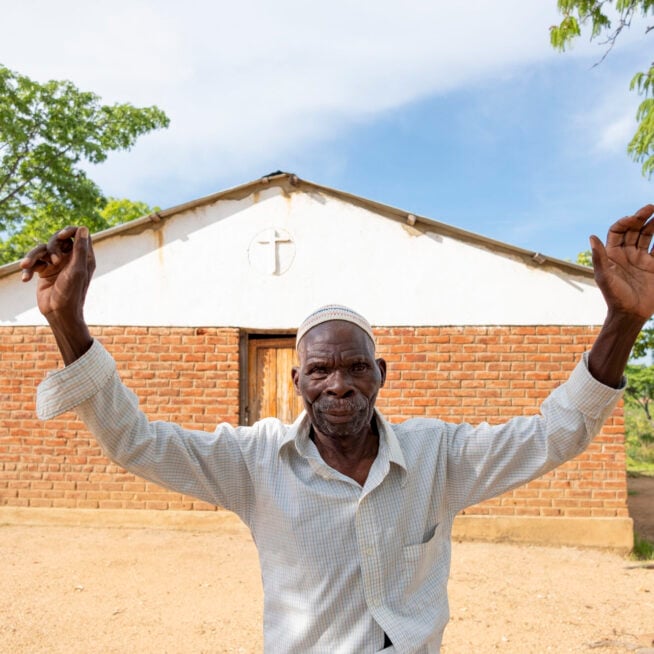 Batwel, wearing a white shirt stands in front of a church building with his hands in the air.