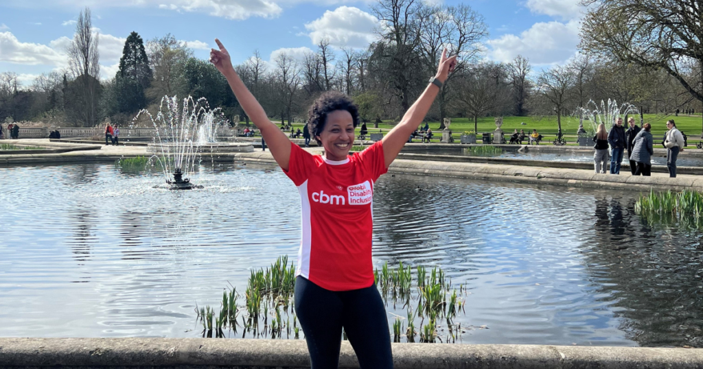 Arwa is wearing a CBM UK running t-shirt. She is standing in front of water. She is training for the London Marathon.