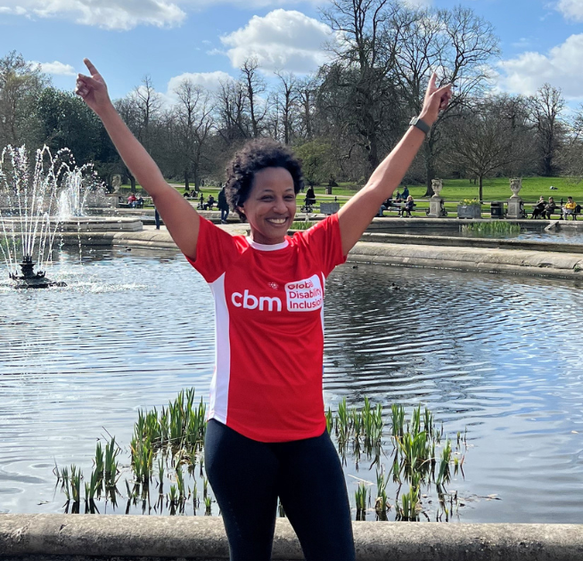 Arwa is wearing a CBM UK running t-shirt. She is standing in front of water. She is training for the London Marathon.