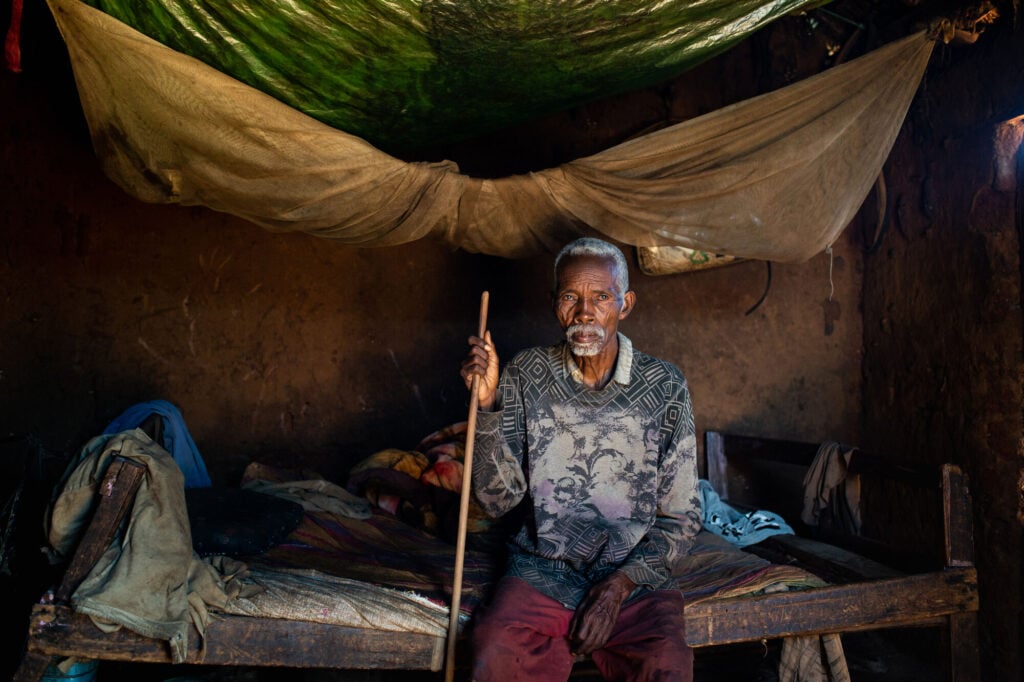 A man sitting indoors on his bed holding his cane.