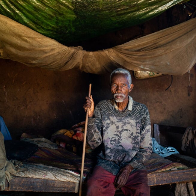 A man sitting indoors on his bed holding his cane.