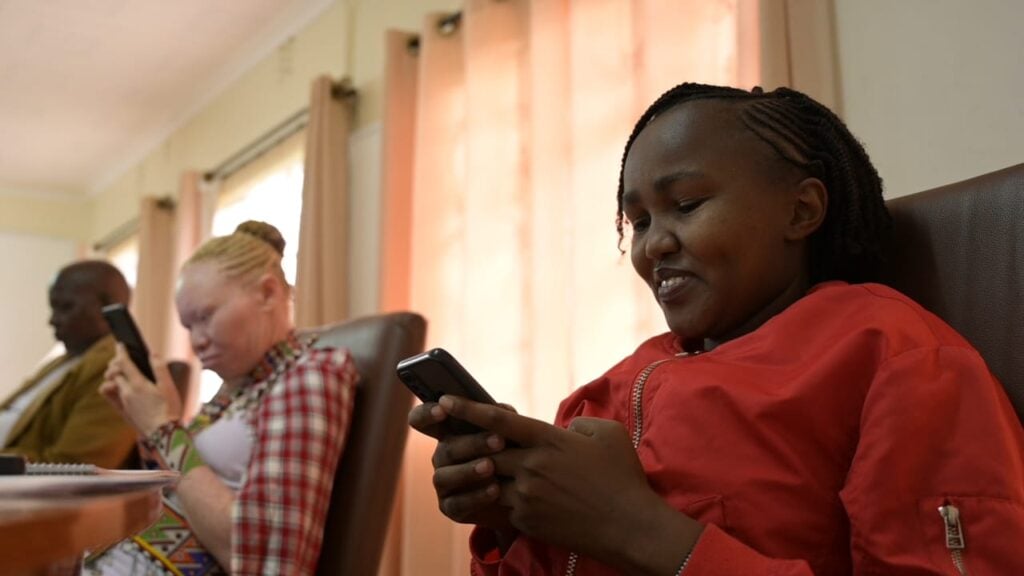 A woman wearing red sits down at a participatory storytelling workshop in Kenya and uses her phone.