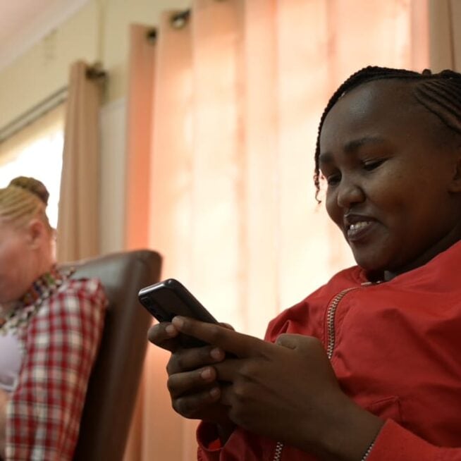 A woman wearing red sits down at a participatory storytelling workshop in Kenya and uses her phone.