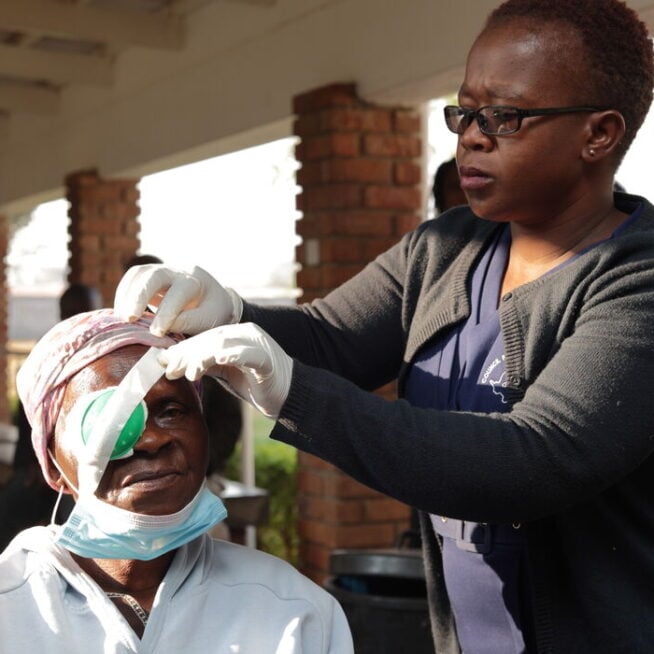 A female healthcare worker adjusts the eye bandage on a elderly male patient who is sitting down.