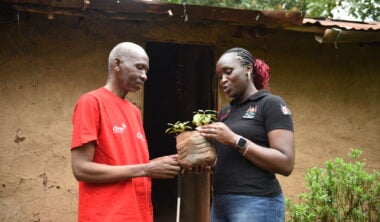 A man and a woman hold a plant pot containing seedlings outside a hut.
