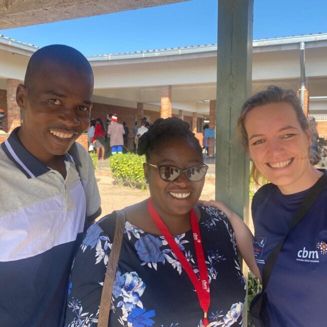 Three people stood outside a hospital in ZImbabwe, smiling into the camera.