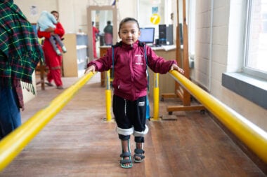 A young girl from Nepal wearing leg braces and learning to walk using yellow barres.