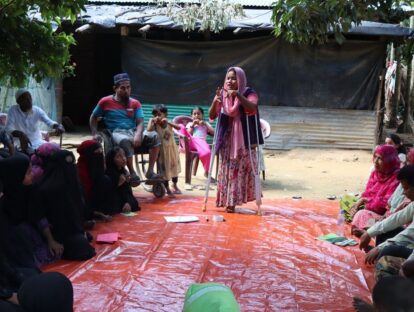 A young woman on crutches stands on a red mat addressing a seated group of people around her.
