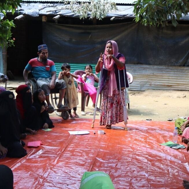 A young woman on crutches stands on a red mat addressing a seated group of people around her.