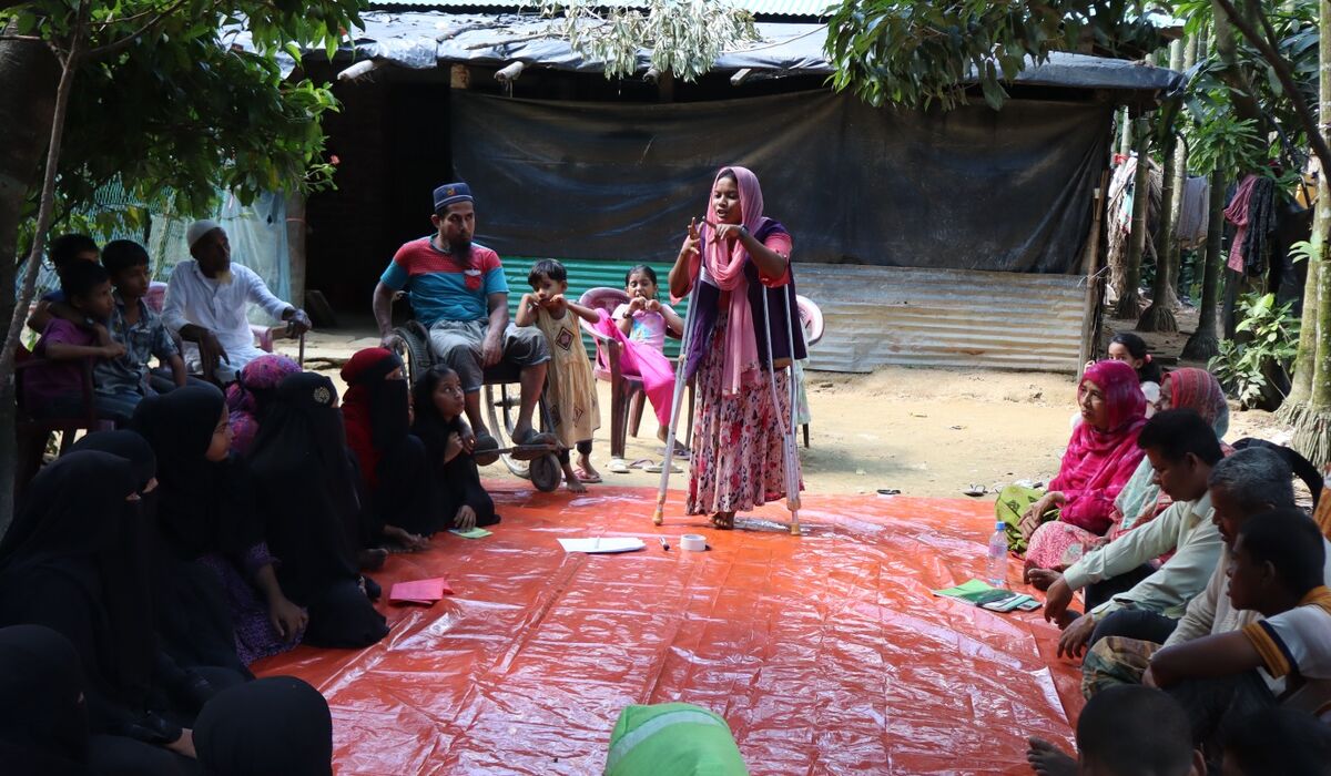 A young woman on crutches stands on a red mat addressing a seated group of people around her.