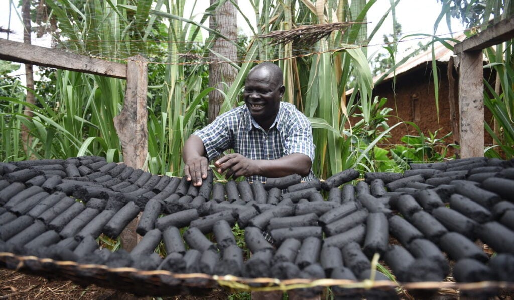 A smiling man stands outdoors at a table arranging a large number of charcoal briquettes.