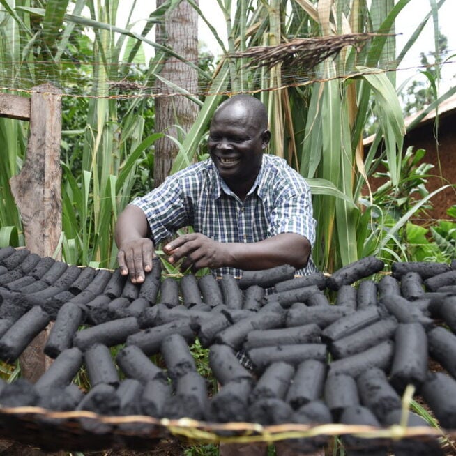 A smiling man stands outdoors at a table arranging a large number of charcoal briquettes.