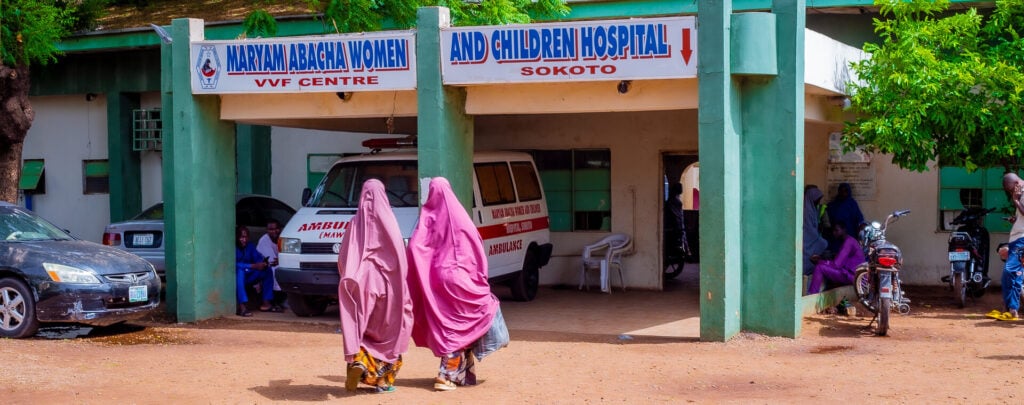 Two women in pink chadors walk across a dusty road towards a building called 'Women and Children Hospital'.