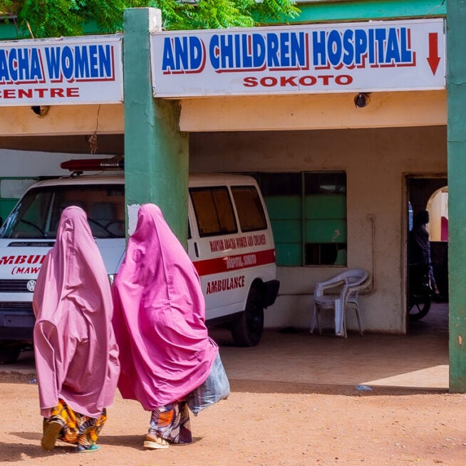 Two women in pink chadors walk across a dusty road towards a building called 'Women and Children Hospital'.
