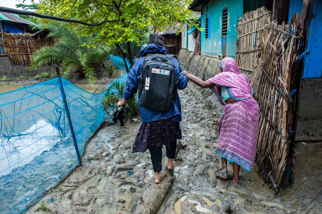 Someone with a rucksack guides a woman wearing purple along a muddy path.