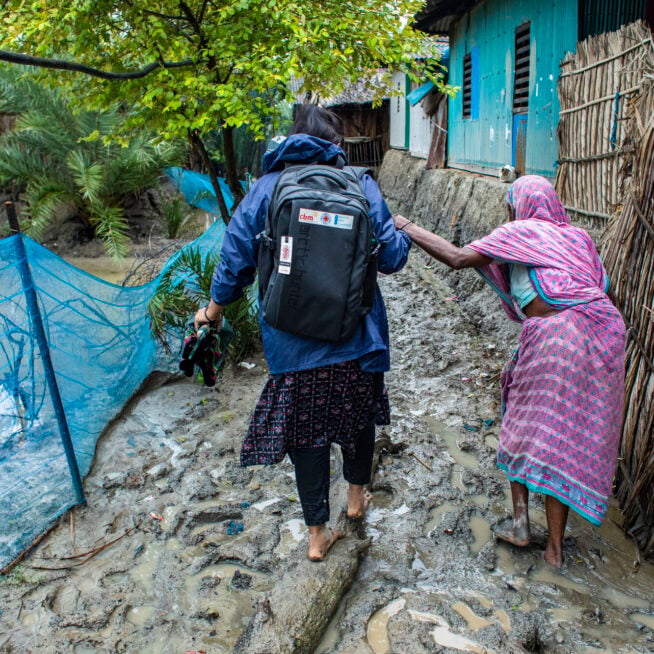 Someone with a rucksack guides a woman wearing purple along a muddy path.