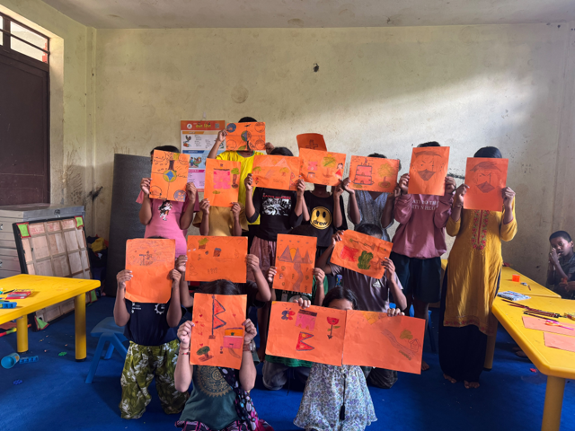 A group of children in a classroom hold up their work on orange sheets of paper.